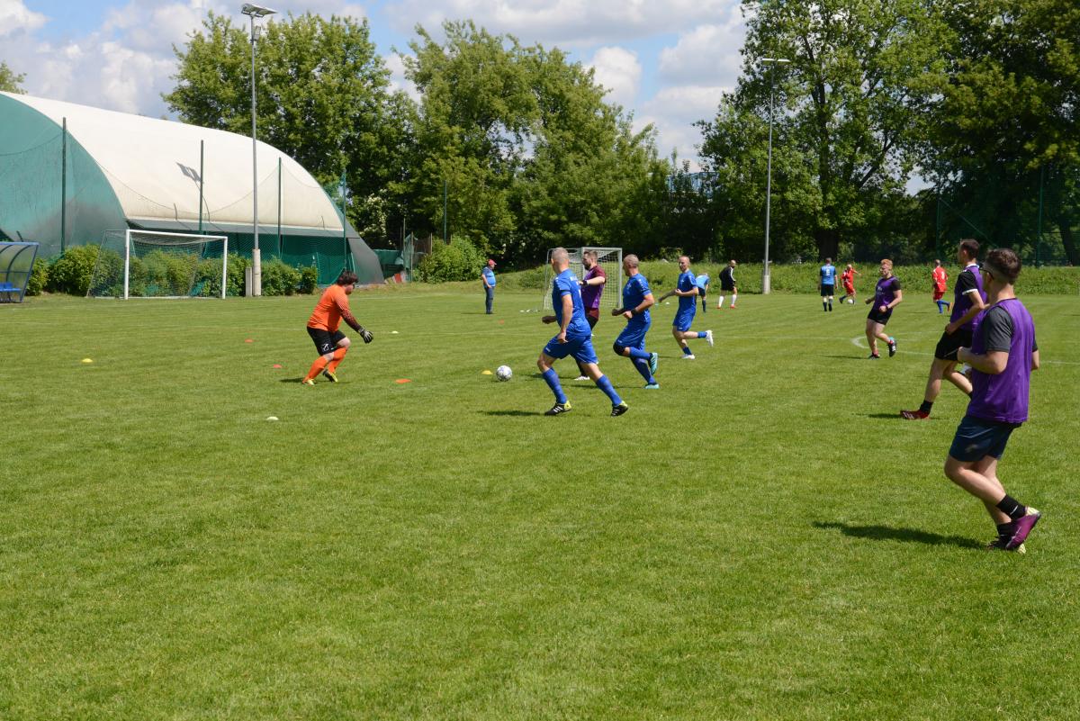 Zdjęcie nr: 16-DSC_3275 przedstawia odbywający się na murawie mecz piłkarski drużyn ubranych w niebieskie stroje oraz w stroje fioletowoczarne. Bramkarz ubrany w strój pomarańczowy podejmuje próbę obrony przed golem. Trzech piłkarz biegnie w kierunku bramki.