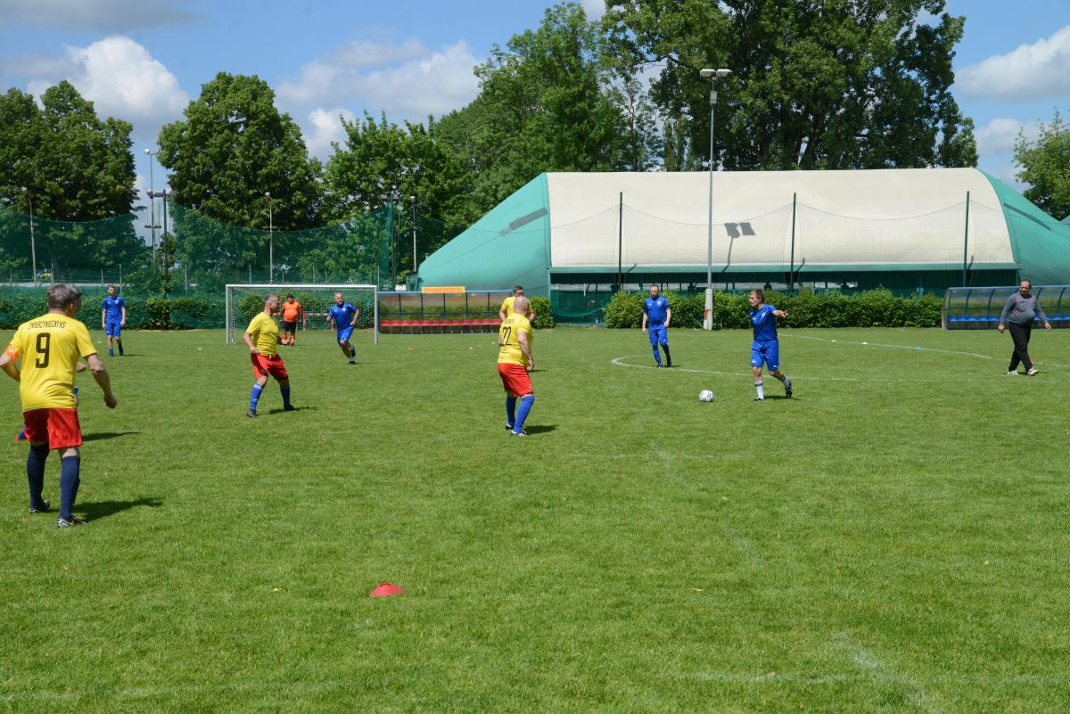 Zdjęcie nr: 19-DSC_3291 przedstawia odbywający się na murawie mecz piłkarski drużyn ubranych w żółtoczerwone stroje oraz w stroje koloru niebieskiego. W tle mecz dogląda sędzia piłkarski. W oddali jest bramkarz ubrany w pomarańczową koszulkę.