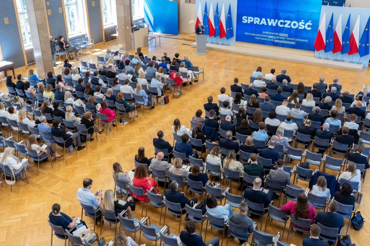 Duża sala konferencyjna wypełniona ludźmi siedzącymi na krzesłach, skierowanymi w stronę sceny. Na scenie znajduje się mównica, przy której stoi prelegent. Za nim widoczne są flagi Polski oraz Unii Europejskiej, a na dużym ekranie wyświetlone jest słowo „SPRAWCZOŚĆ” oraz tekst w języku polskim. Uczestnicy są elegancko ubrani, a sala jest jasna, z drewnianą podłogą i dużymi oknami po lewej stronie.