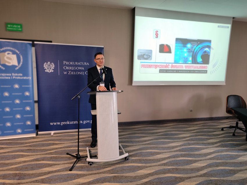 Na zdjęciu znajduje się mężczyzna w garniturze, stojący za mównicą i przemawiający do publiczności. Przed nim ustawiony jest mikrofon na statywie. W tle po lewej stronie widoczne są dwa niebieskie banery – jeden z logo i napisem „Prokuratura Okręgowa w Zielonej Górze”, a drugi z logotypem Krajowej Szkoły Sądownictwa i Prokuratury. Po prawej stronie na ścianie wyświetlona jest prezentacja multimedialna. Na slajdzie widoczne są grafiki związane z technologią oraz napis: „Przestępczość świata wirtualnego – czyli zagrożenia i narzędzia ich zwalczania”. Pomieszczenie jest jasne, z dywanem w faliste wzory i kilkoma krzesłami ustawionymi przy ścianie.