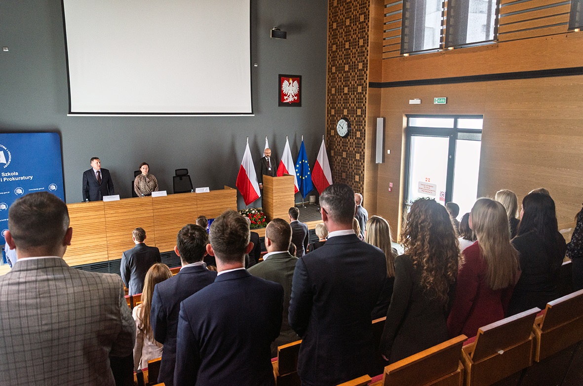 Wnętrze nowoczesnej sali konferencyjnej z drewnianymi ścianami i dużym oknem po prawej stronie, przez które wpada światło dzienne. W pierwszym planie widoczni są uczestnicy wydarzenia, którzy stoją w rzędach skierowani w stronę podwyższenia. Uczestnicy ubrani są elegancko, w garnitury i formalne stroje. Na podwyższeniu znajduje się długi drewniany stół, za którym stoją dwie osoby, a trzecia osoba znajduje się za mównicą. W tle ustawione są cztery flagi Polski, flaga Unii Europejskiej oraz flaga instytucji. Na ścianie wisi godło Polski oraz zegar. Po lewej stronie widoczna jest niebieska plansza z logo i nazwą „Krajowa Szkoła Sądownictwa i Prokuratury”. Atmosfera jest oficjalna i uroczysta.