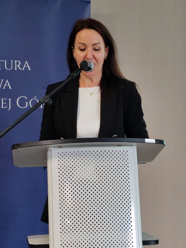 Na zdjęciu widać kobietę stojącą za mównicą podczas oficjalnego wydarzenia. Ubrana jest elegancko – ma na sobie czarną marynarkę i białą bluzkę. Przed nią znajduje się mikrofon na statywie, skierowany w jej stronę. W tle widoczny jest niebieski baner z napisem „Prokuratura Okręgowa w Zielonej Górze” oraz godłem państwowym. 