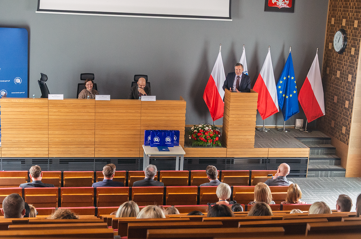 Widok sali konferencyjnej z drewnianymi ławkami, w której odbywa się oficjalne wydarzenie. Na pierwszym planie siedzi publiczność, a na podwyższeniu zasiadają dwie osoby za dużym stołem. Po prawej stronie mężczyzna w garniturze przemawia z mównicy. W tle widoczne są cztery flagi Polski oraz flaga Unii Europejskiej, a także kwiaty ustawione na podłodze. Na ścianie znajduje się zegar i godło Polski.