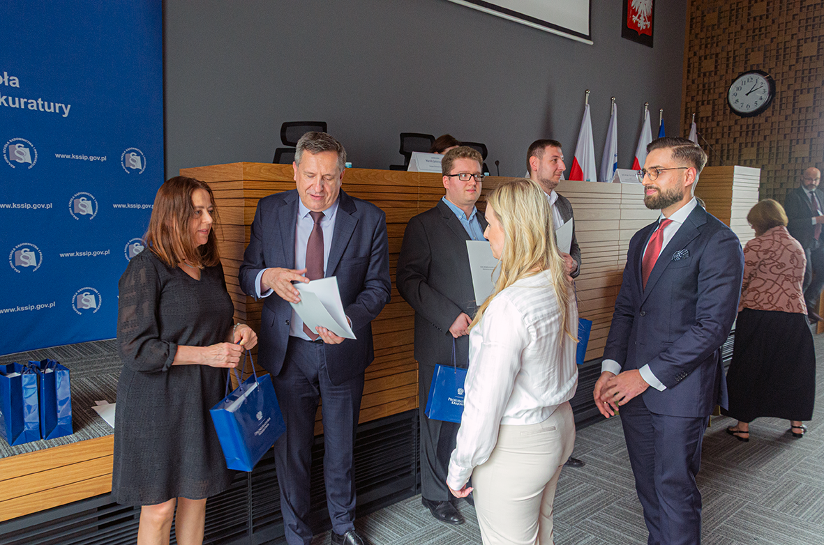 Grupa osób w eleganckich strojach uczestniczy w uroczystym wręczeniu gratulacji w pobliżu sceny w sali konferencyjnej. Kilkoro uczestników otrzymuje oficjalne dokumenty oraz niebieskie torby z logo instytucji od przedstawicieli organizatora. W tle znajduje się niebieska ścianka z nazwą organizatora oraz fragment sceny z drewnianą zabudową i flagami Polski, Unii Europejskiej oraz instytucji. Na ścianie widoczny jest zegar oraz godło Polski. Po prawej stronie, w oddali, widać kolejne osoby przemieszczające się po sali. 
