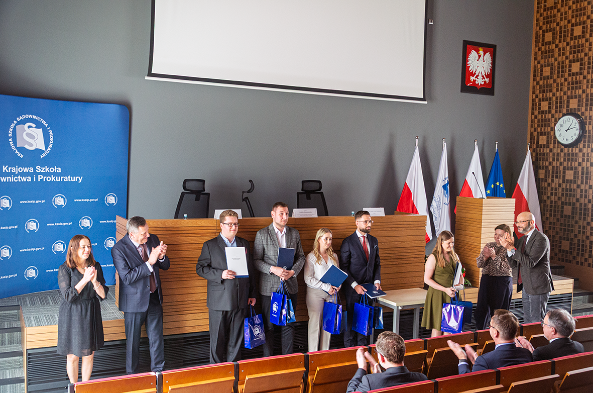 Grupa osób stoi na scenie w sali konferencyjnej podczas oficjalnej uroczystości. Kilka osób trzyma w rękach dyplomy oraz niebieskie torby z logo instytucji. W tle widoczne są flagi Polski, Unii Europejskiej oraz Krajowej Szkoły Sądownictwa i Prokuratury. Na ścianie wisi godło Polski i zegar. Przed sceną siedzi publiczność, która bije brawo.