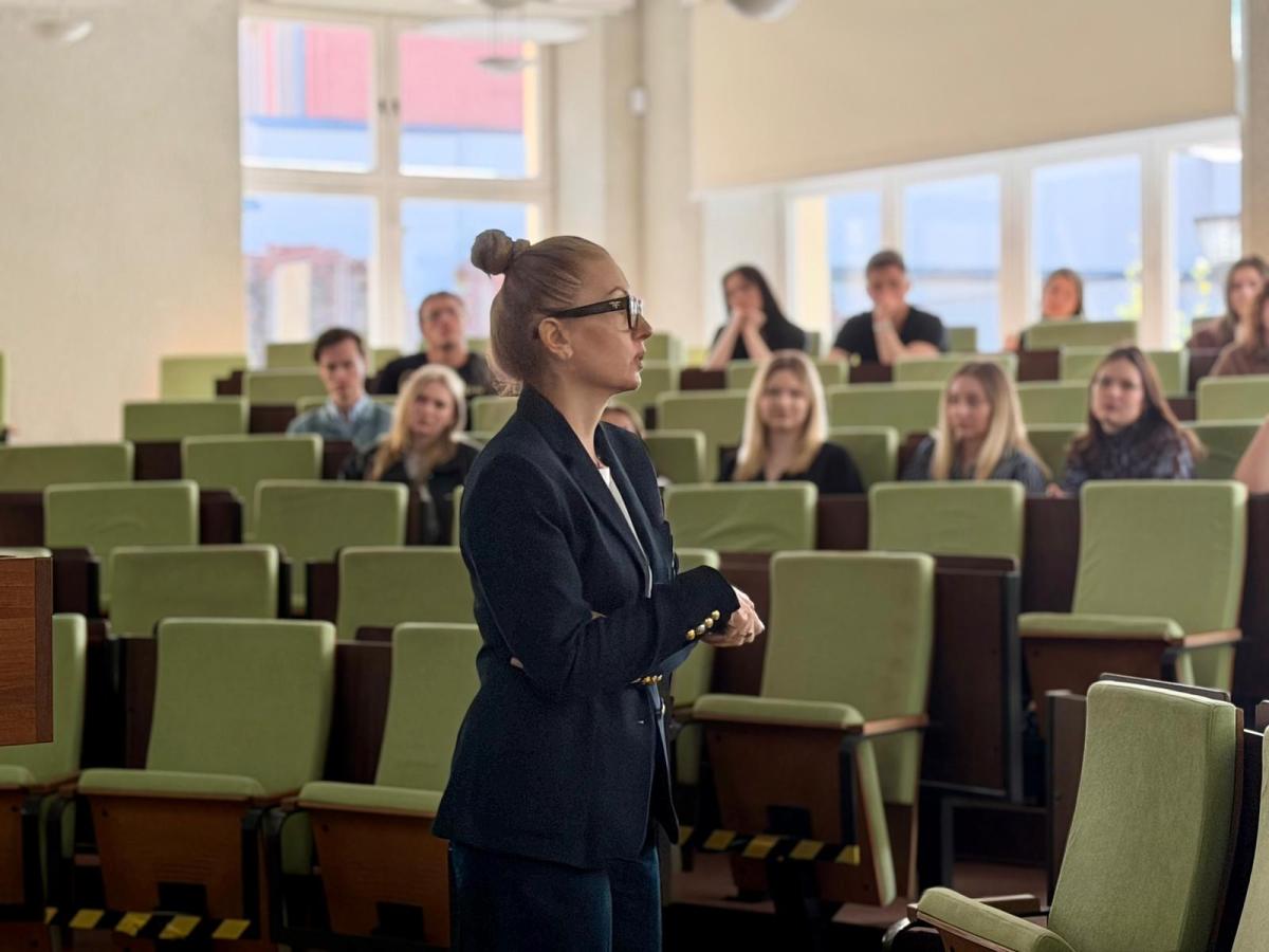 Kobieta w ciemnym garniturze stoi bokiem w sali wykładowej i prowadzi prezentację dla grupy studentów siedzących w rzędach zielonych foteli. W tle widoczne są duże okna oraz uczestnicy słuchający wystąpienia. Sala jest jasna i przestronna.