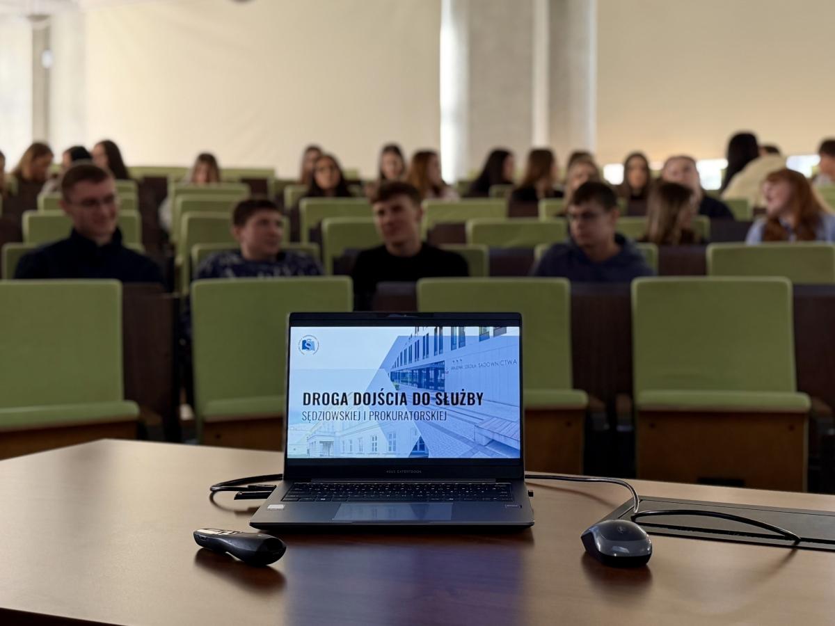 W sali wykładowej stoi pulpit z otwartym laptopem, na którego ekranie wyświetlona jest prezentacja zatytułowana „Droga dojścia do służby sędziowskiej i prokuratorskiej”. Obok laptopa leży mysz komputerowa i pilot. W tle widać rzędy zielonych foteli audytoryjnych, na których siedzi grupa osób oczekujących na rozpoczęcie prezentacji. Pomieszczenie jest jasne, z zasłoniętymi dużymi oknami.