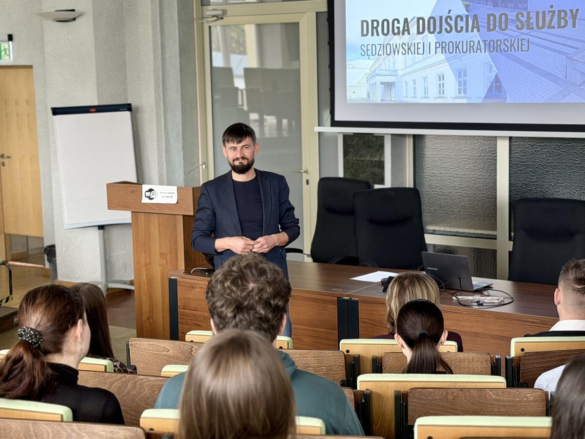 Mężczyzna prowadzi prezentację w sali szkoleniowej, stojąc przed stołem i mównicą. Ubrany jest w ciemną marynarkę i czarną koszulkę, ma krótkie włosy i brodę. Przed nim siedzi grupa uczestników, widocznych od tyłu, zwróconych w jego stronę. W tle znajduje się ekran z prezentacją zatytułowaną „Droga dojścia do służby sędziowskiej i prokuratorskiej”, a także drzwi i elementy wyposażenia sali, w tym flipchart i krzesła. Atmosfera spotkania ma charakter formalny i edukacyjny. 