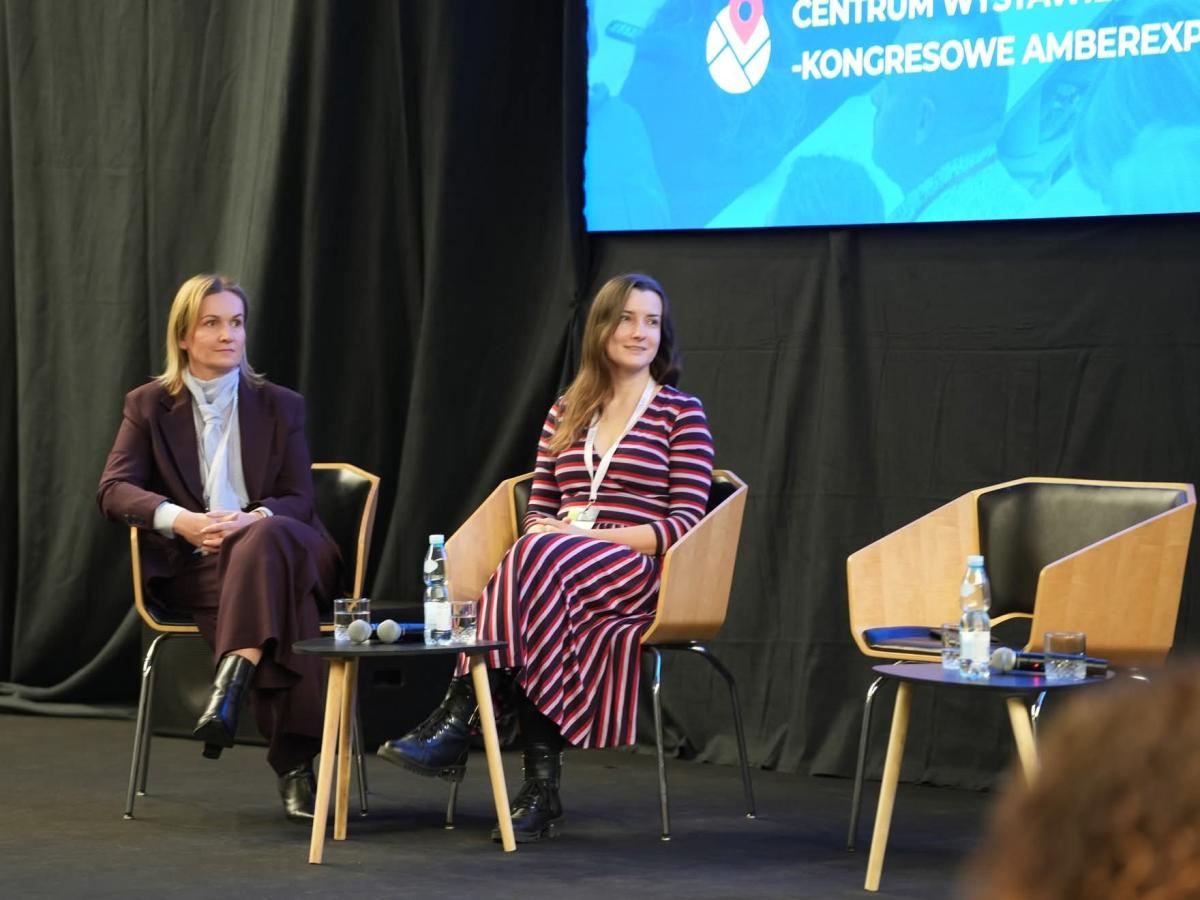 Dwie kobiety siedzą na scenie podczas panelu dyskusyjnego w centrum konferencyjnym AmberExpo. Przed nimi stoją stoliki z mikrofonami, szklankami i butelkami wody. W tle widoczny jest czarny materiał sceniczny i fragment niebieskiego ekranu z logo Centrum Wystawienniczo-Kongresowego AmberExpo.