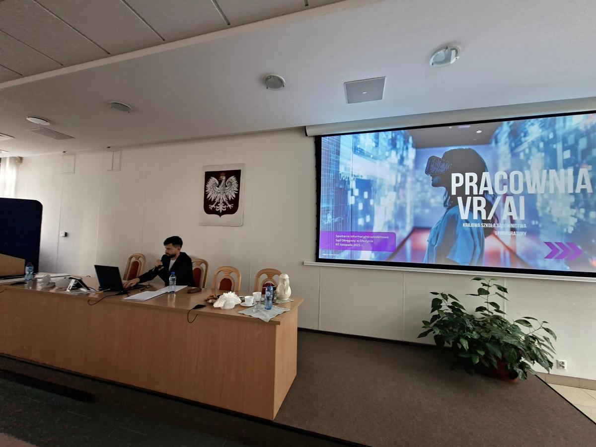 Sala konferencyjna z dużym ekranem, na którym widoczny jest napis Pracownia VR/AI oraz grafika osoby w goglach VR w futurystycznym otoczeniu. Prelegent siedzi przy stole z laptopem, obok stoją krzesła. Na ścianie wisi godło Polski, a pod ekranem znajduje się roślina w doniczce.