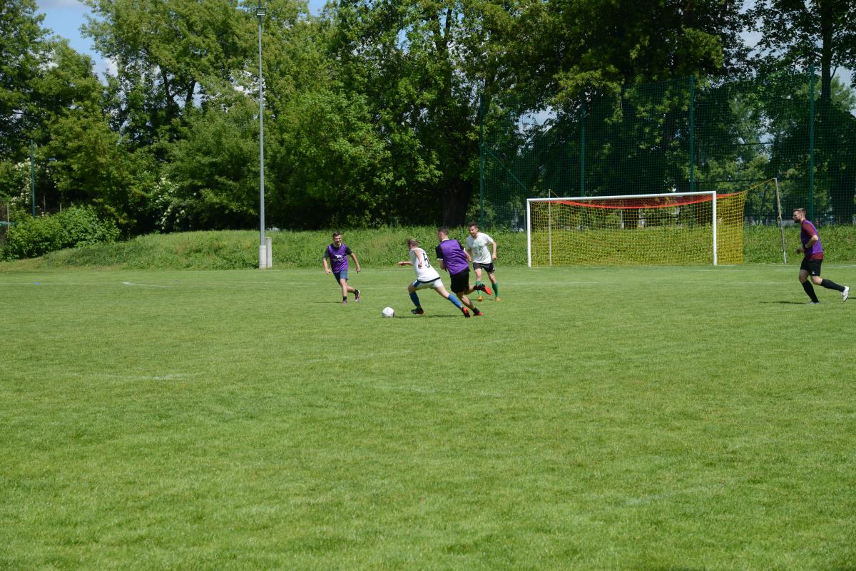 Zdjęcie nr: 20-DSC_3294 przedstawia odbywający się na murawie mecz piłkarski drużyn ubranych w jasnoczarne stroje oraz w stroje fioletowoczarne. Zawodnicy biegają po boisku za  piłką.