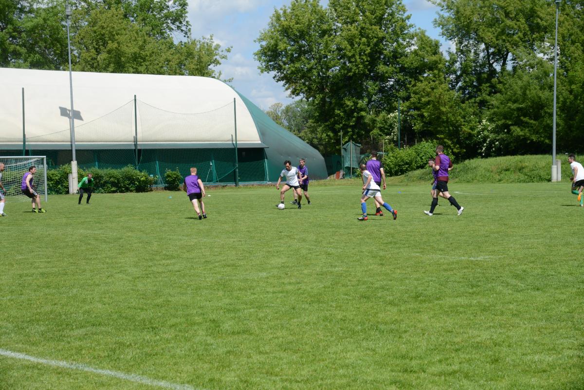 Zdjęcie nr: 21-DSC_3295 przedstawia odbywający się na murawie mecz piłkarski drużyn ubranych w jasnoczarne stroje oraz w stroje fioletowoczarne. Zawodnicy biegają po boisku za  piłką.