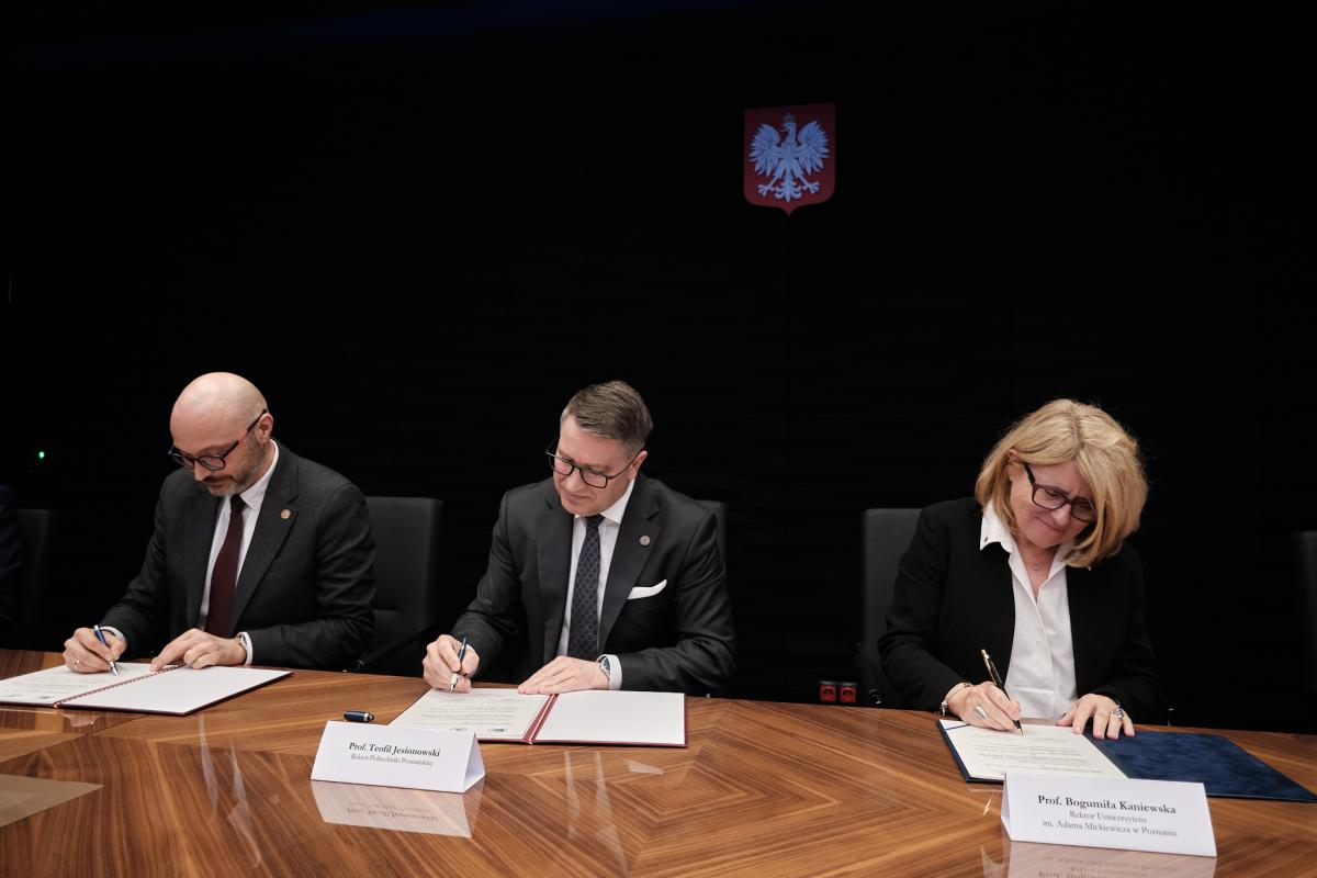 Na zdjęciu widoczne są trzy osoby: dwóch mężczyzn oraz kobieta, siedzące przy stole, skupione na podpisywaniu dokumentów. Ubrani są formalnie, co podkreśla oficjalny charakter sytuacji. W tle znajduje się czarne tło oraz widoczne godło, nadające całości powagi i urzędowego kontekstu.
