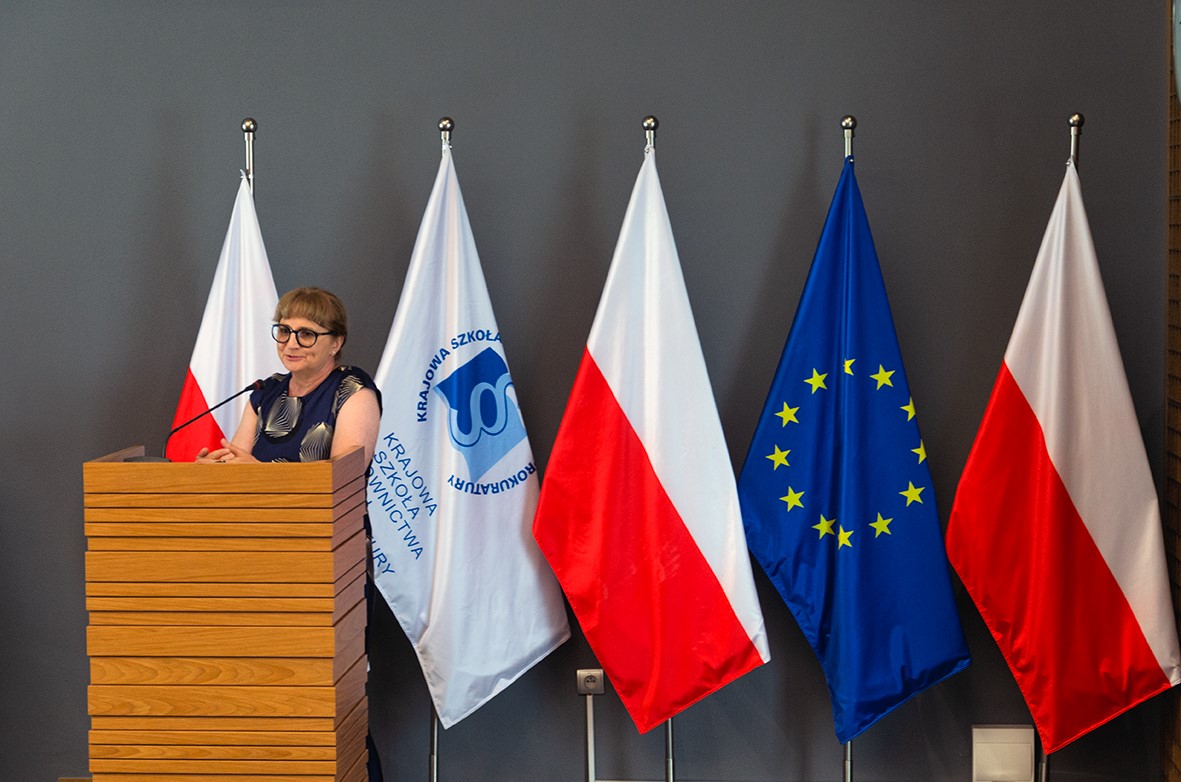 Kobieta w okularach stoi za drewnianą mównicą i przemawia do mikrofonu. W tle znajdują się cztery flagi: dwie biało-czerwone flagi Polski, jedna flaga Unii Europejskiej oraz biała flaga z niebieskim logo Krajowej Szkoły Sądownictwa i Prokuratury. Ściana jest szara.