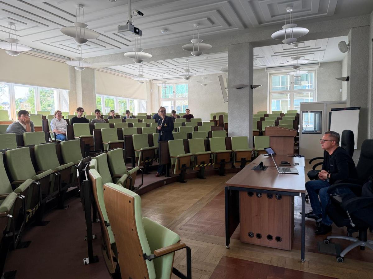 Widok na dużą salę wykładową z rzędami zielonych foteli. Przy biurku z komputerem siedzi mężczyzna prowadzący spotkanie, a kilka osób uczestniczy w zajęciach z różnych miejsc na sali. W centralnej części stoi kobieta prowadząca rozmowę lub prezentację. Wnętrze jest nowoczesne i dobrze oświetlone.