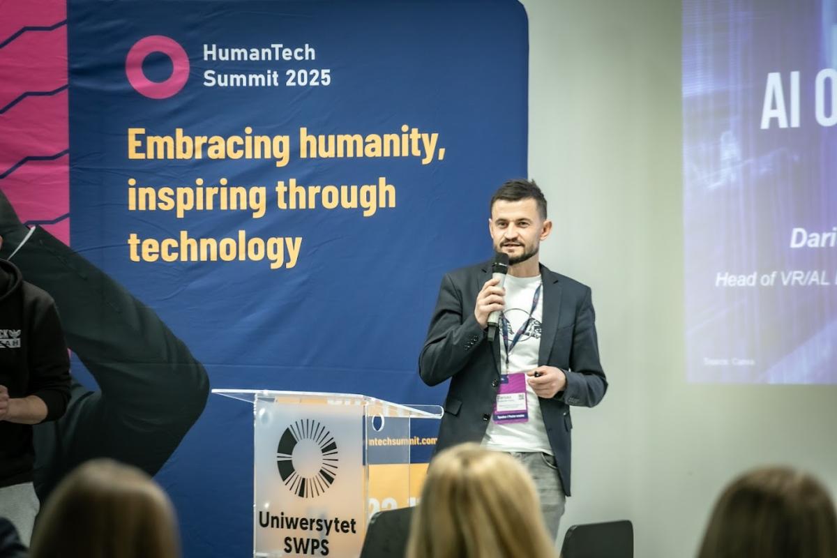 Mężczyzna w marynarce stoi przy mównicy z logo Uniwersytetu SWPS i mówi do mikrofonu. Za nim znajduje się duży niebiesko-różowy baner konferencyjny z napisem „HumanTech Summit 2025. Embracing humanity, inspiring through technology”. Przed nim siedzą słuchacze.