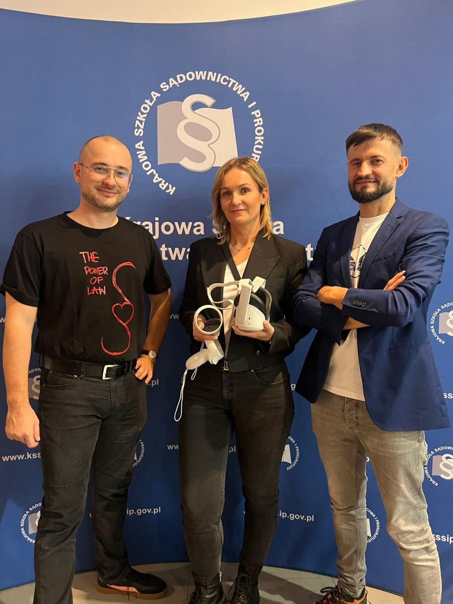 Trzy osoby stoją obok siebie przed niebieską ścianką z logo Krajowej Szkoły Sądownictwa i Prokuratury. Wszyscy patrzą w obiektyw i uśmiechają się. Osoba w środku trzyma gogle VR oraz kontrolery, symbolizujące nowoczesne narzędzia edukacyjne wykorzystywane w szkoleniach sędziów i prokuratorów. 