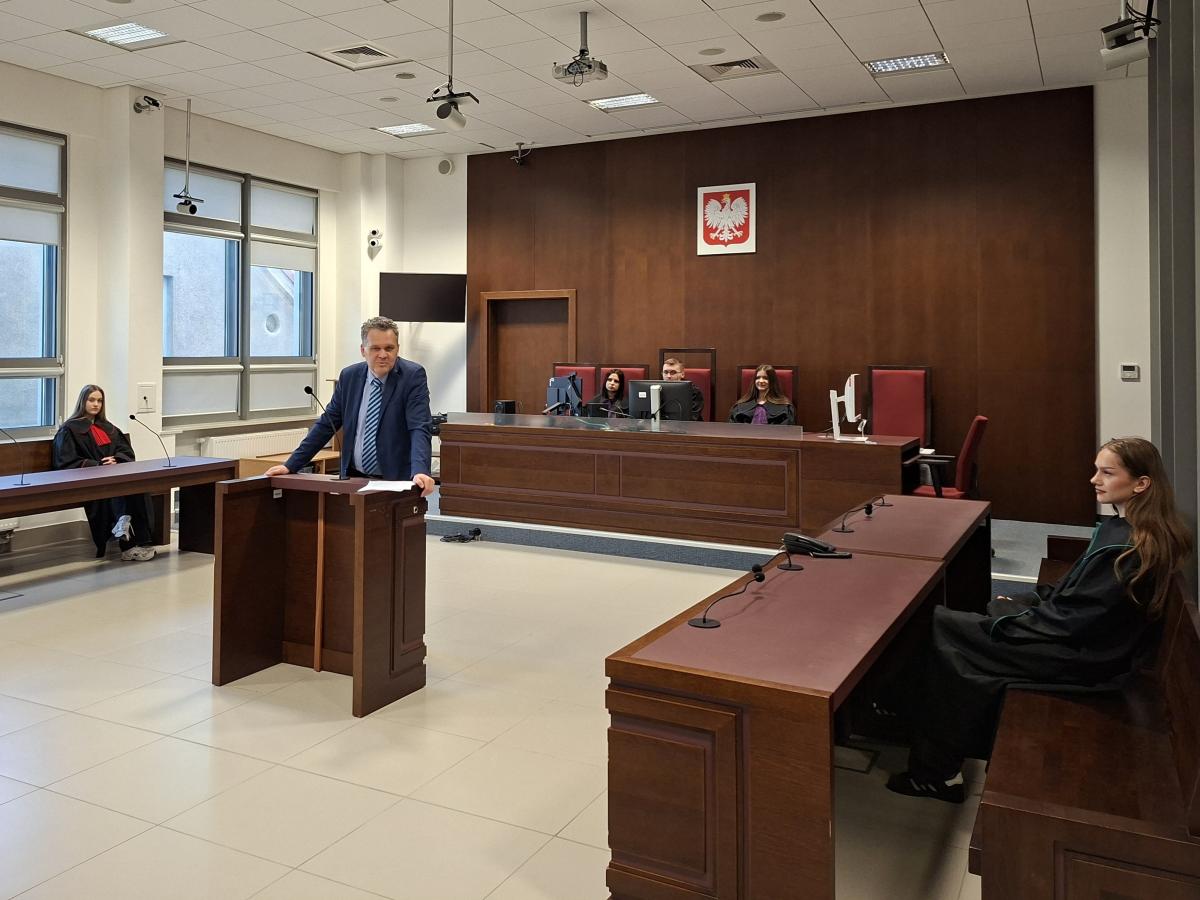 W nowoczesnej sali sądowej znajdują się rzędy stołów i krzeseł wykonanych z ciemnego drewna. Na ścianie z tyłu zamontowane jest godło państwowe. Trzy osoby w togach z fioletowym żabotem siedzą przy stole sędziowskim, a jedna osoba stoi przy mównicy pośrodku sali. Po bokach widoczne są miejsca do siedzenia dla uczestników rozprawy, przy których siedzą dwie młode kobiety w togach. W pomieszczeniu jest jasne oświetlenie i duże okna z roletami.