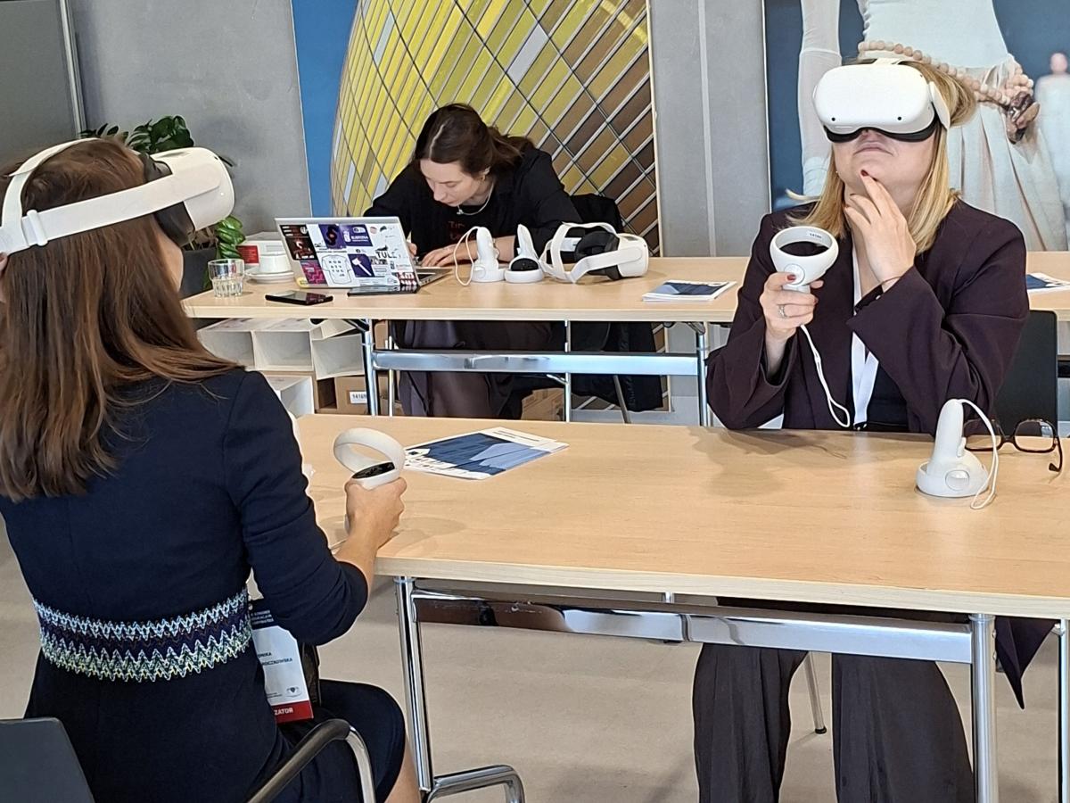 Uczestniczki szkolenia siedzą przy stołach i korzystają z gogli rzeczywistości wirtualnej. Każda trzyma kontrolery VR. W tle inna osoba pracuje przy laptopie. Na stołach leżą ulotki Krajowej Szkoły Sądownictwa i Prokuratury.