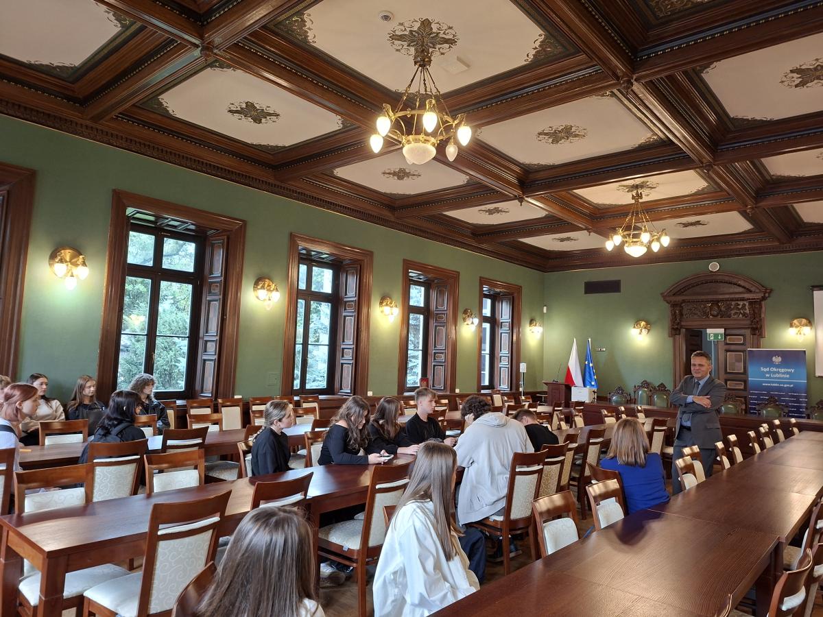 Sala w zabytkowym budynku sądu, z zielonymi ścianami, drewnianym sufitem kasetonowym i dużymi oknami. Przy długich stołach siedzi grupa młodych osób, a z przodu sali stoi mężczyzna w garniturze. Obok widoczne są flagi Polski i Unii Europejskiej oraz baner Sądu Okręgowego w Lublinie.