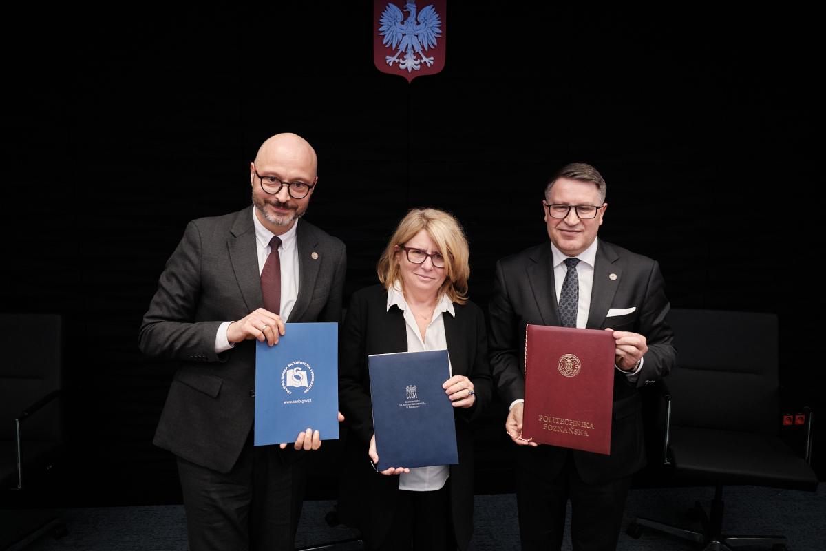 Na zdjęciu widoczne są trzy osoby stojące obok siebie, trzymające w rękach teczki z podpisanymi dokumentami. Ubrane są w formalne stroje, co podkreśla oficjalny charakter wydarzenia. Tłem jest czarna ściana z widocznym godłem, nadająca całej scenie powagę i uroczysty klimat. Za mężczyzna widać czarny fotel.