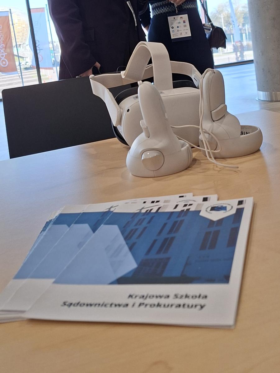 Na stole leżą białe gogle rzeczywistości wirtualnej z dwoma kontrolerami oraz ulotki Krajowej Szkoły Sądownictwa i Prokuratury. W tle widać dwie osoby w formalnych ubraniach rozmawiające w jasnym pomieszczeniu konferencyjnym.