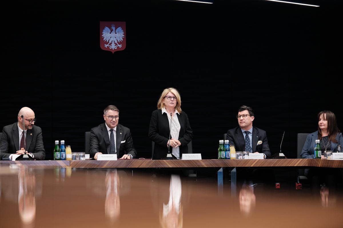 Na zdjęciu widoczny jest długi blat stołu, za którym znajduje się pięć osób – trzech mężczyzn i dwie kobiety, wszyscy ubrani formalnie. Jedna z kobiet stoi, trzymając przed sobą złożone ręce i przemawia do mikrofonu. Na stole znajdują się napoje oraz tabliczki z nazwiskami. W tle widać czarne tło z godłem, co nadaje scenie oficjalny i uroczysty charakter.