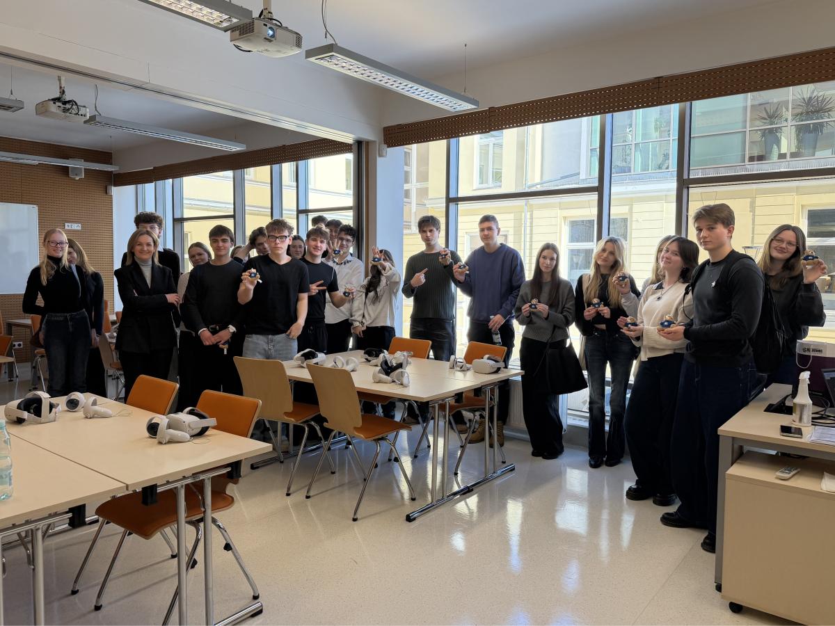 Wizyta w Pracowni VR/AI; Grupa młodych osób wraz z dorosłą kobietą stoi w jasnej sali przy stolikach, na których leżą gogle wirtualnej rzeczywistości. Uczestnicy trzymają w dłoniach małe pluszowe misie i patrzą w stronę aparatu. W pomieszczeniu znajdują się krzesła, rzutnik sufitowy oraz duże okna z widokiem na budynek po drugiej stronie dziedzińca.
