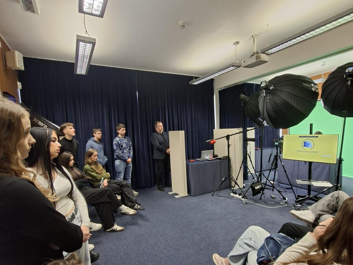 Grupa młodych osób słucha prowadzącego w sali wyposażonej w sprzęt do nagrań wideo. W pomieszczeniu znajdują się duże lampy studyjne, statywy z kamerami oraz zielone tło do nagrań. Część uczestników stoi, część siedzi. Okna zasłonięte są aksamitnymi zasłonami w kolorze granatowym.