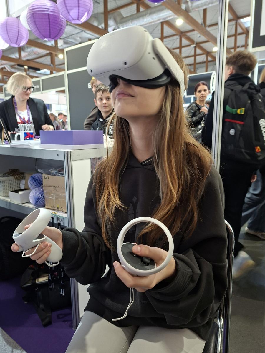 Młoda kobieta w czarnej bluzie siedzi na krześle i korzysta z gogli wirtualnej rzeczywistości. W dłoniach trzyma kontrolery. Za nią widoczne jest stoisko targowe, udekorowane fioletowymi lampionami, obok którego przechodzą młodzi ludzie.