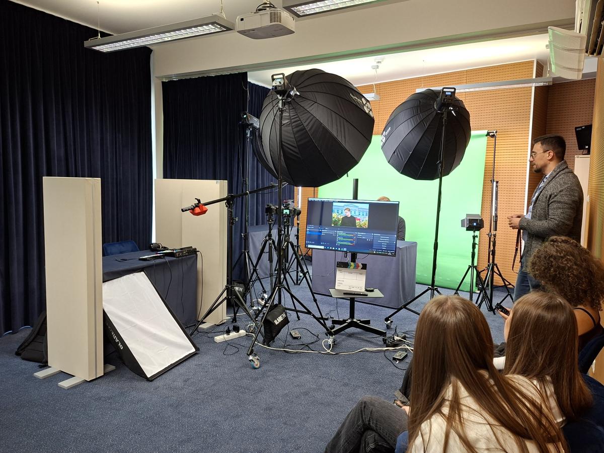 Studio nagraniowe z profesjonalnym sprzętem do produkcji wideo. W centralnej części znajduje się monitor wyświetlający podgląd nagrania z efektem greenscreenu – widać na nim osobę na tle budynku z ogrodem. Obok monitora stoi mężczyzna, prowadzący warsztaty. Na środku pomieszczenia ustawione są statywy z dużymi czarnymi lampami studyjnymi oraz mikrofon kierunkowy. W tle widoczna jest zielona płachta greenscreenu. Po prawej stronie siedzą trzy młode kobiety, które uważnie obserwują wydarzenie. 