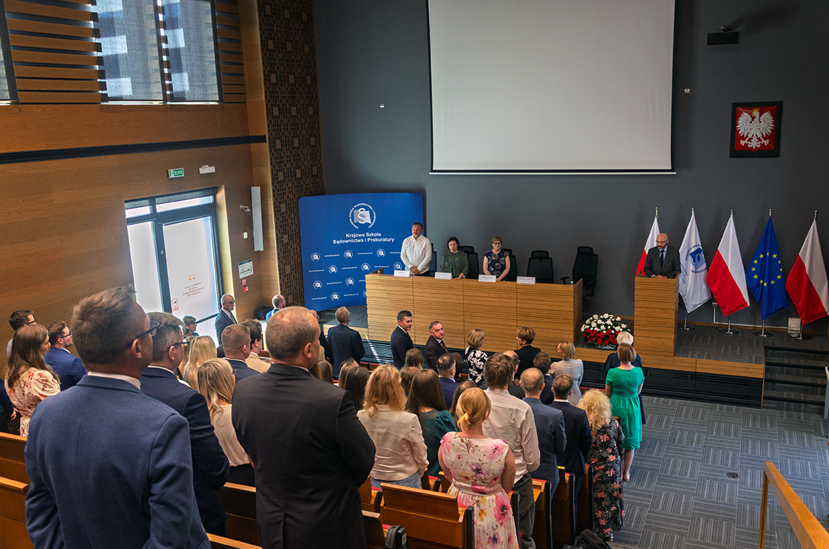 Sala wykładowa z drewnianymi ścianami i dużymi oknami po lewej stronie. W pierwszym planie widoczni są uczestnicy wydarzenia stojący w rzędach czerwonych foteli. Na podwyższeniu z przodu sali znajduje się panel z czterema osobami siedzącymi za długim, drewnianym stołem oraz mówca przy mównicy. Za mównicą stoją cztery flagi: dwie polskie, flaga Unii Europejskiej oraz flaga instytucji. Na ścianie wisi godło Polski. Po lewej stronie stoi niebieska tablica z napisem „Krajowa Szkoła Sądownictwa i Prokuratury”.