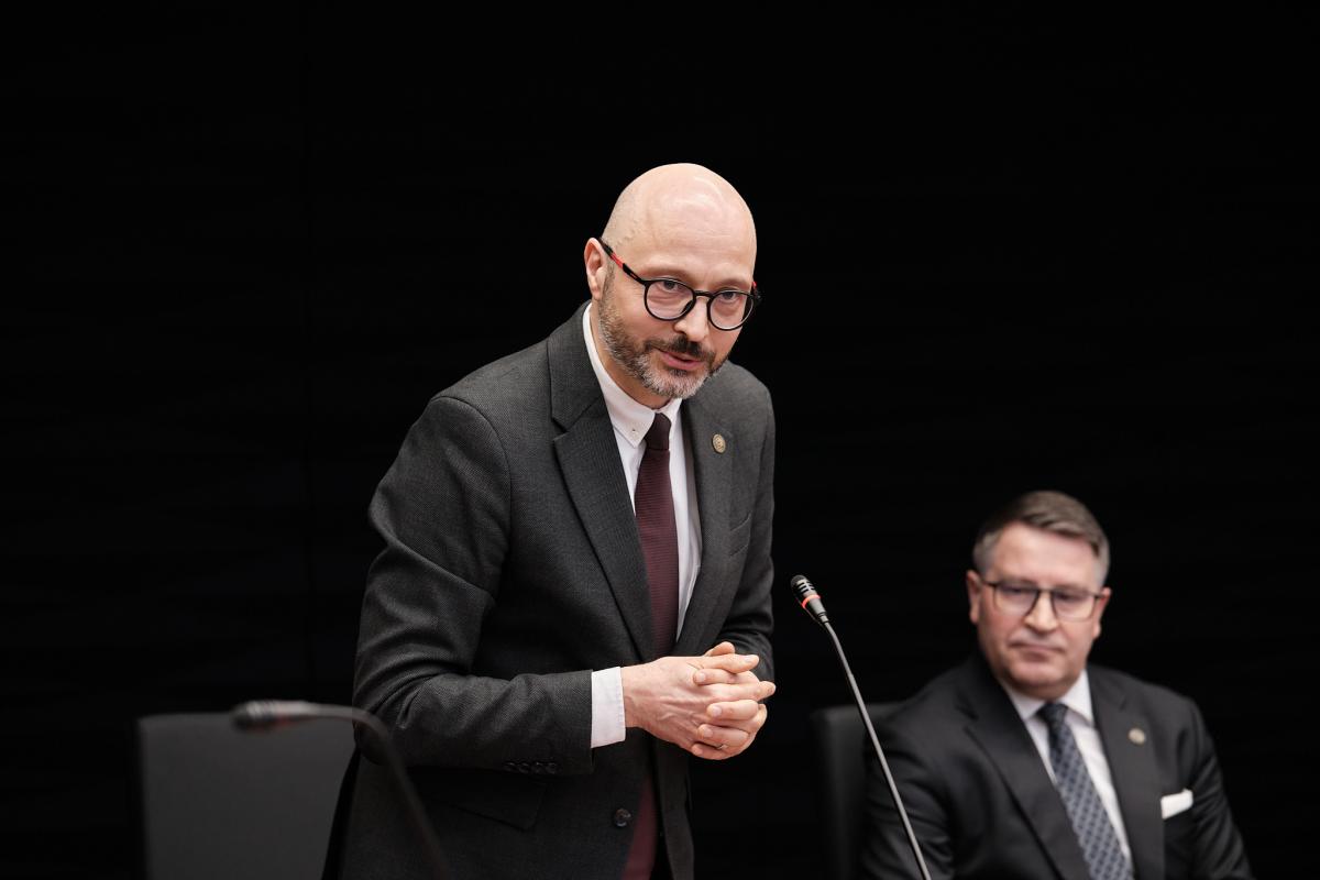 Na zdjęciu Dyrektor Krajowej Szkoły Sądownictwa i Prokuratury stoi i przemawia do mikrofonu. Ubrany jest w elegancki garnitur, ma złożone przed sobą ręce i lekko się uśmiecha. Obok niego siedzi mężczyzna również ubrany formalnie. W tle widoczne jest czarne tło, które podkreśla oficjalny charakter sytuacji.