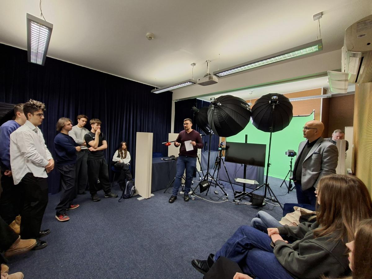 Wizyta w Studiu nagrań multimedialnych; Grupa młodych osób słucha prowadzącego w sali wyposażonej w sprzęt do nagrań wideo. W pomieszczeniu znajdują się duże lampy studyjne, statywy z kamerami oraz zielone tło do nagrań. Część uczestników stoi, część siedzi, a prowadzący trzyma kartkę i mówi do grupy.