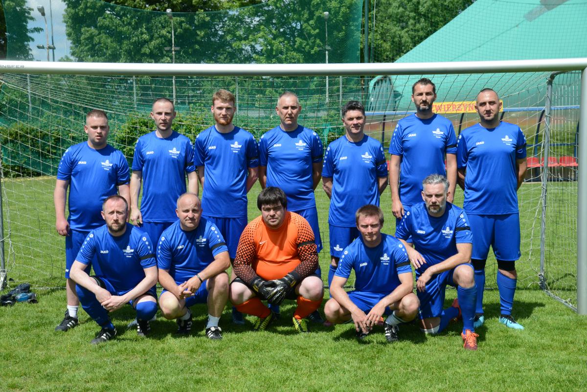 Zdjęcie nr: 6-DSC_3281 przedstawia skład męskiej drużyny piłkarskiej, składającej się z dwunastu zawodników, na tle bramki piłkarskiej, na murawie, jedenastu z nich ubranych jest strój piłkarski koloru  niebieskiego a jeden w strój bramkarski koloru pomarańczowego. Siedmiu mężczyzn stoi, pozostali spoczywają (kucają) na trawie.
