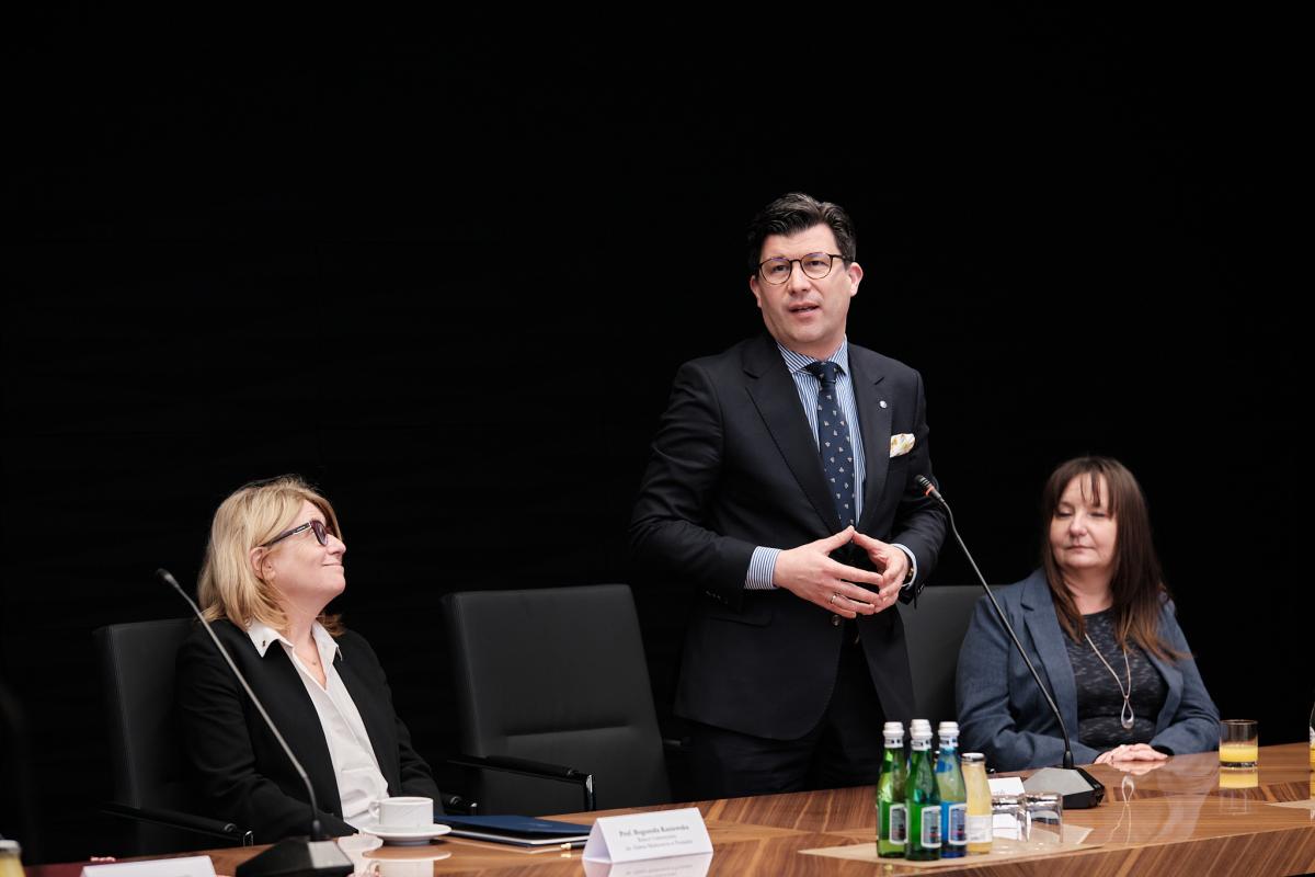 Na zdjęciu widoczne są trzy osoby – dwie kobiety siedzą, a pomiędzy nimi stoi mężczyzna przemawiający do mikrofonu. Ubrany jest w ciemny garnitur, koszulę w paski oraz wzorzysty krawat. Na stole przed nimi znajdują się woda, szklanki, filiżanki oraz tabliczki z nazwiskami. Kobiety słuchają uważnie i są uśmiechnięte. W tle widoczne jest jednolite czarne tło, podkreślające oficjalny charakter wydarzenia.