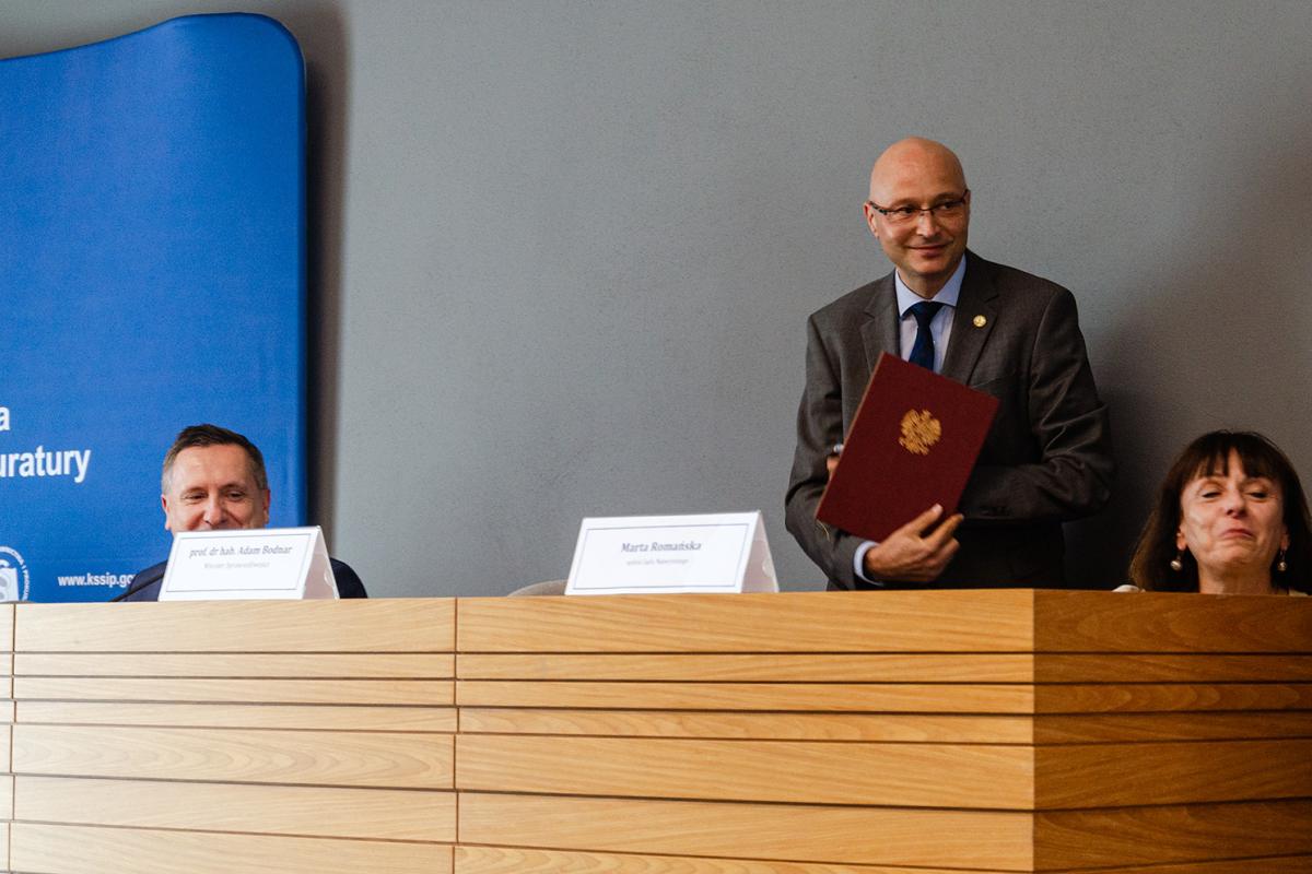 Przedstawia stojącego za katedrą Dyrektora Prof. dr. hab. Piotra Girdwoynia z okładką zawierającą gratulacje objęcia stanowiska. Obok widać Sędzię Sądu Najwyższego Martę Romańską oraz zastępcę Prokuratora Krajowego Marka Jamrogowicza, za którym znajduje się fragment niebieskiej ścianki KSSiP. Zdjęcie wykonano na tle szarej ściany. 