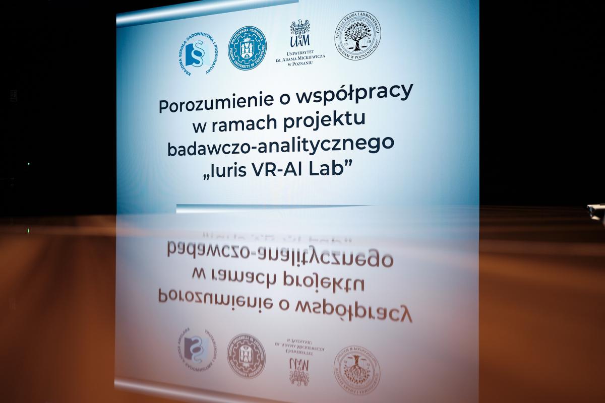 Na zdjęciu widoczny jest wyświetlany slajd zatytułowany Porozumienie o współpracy w ramach projektu badawczo-analitycznego „luris VR-AI Lab”. Nad tytułem znajdują się logotypy KSSiP, Uniwersytetu im. Adama Mickiewicza w Poznaniu, Wydziału Prawa i Administracji UAM w Poznaniu oraz Politechniki Poznańskiej. Obraz slajdu odbija się w powierzchni stołu, a całość utrzymana jest na tle czarnego tła, co podkreśla oficjalny charakter wydarzenia.