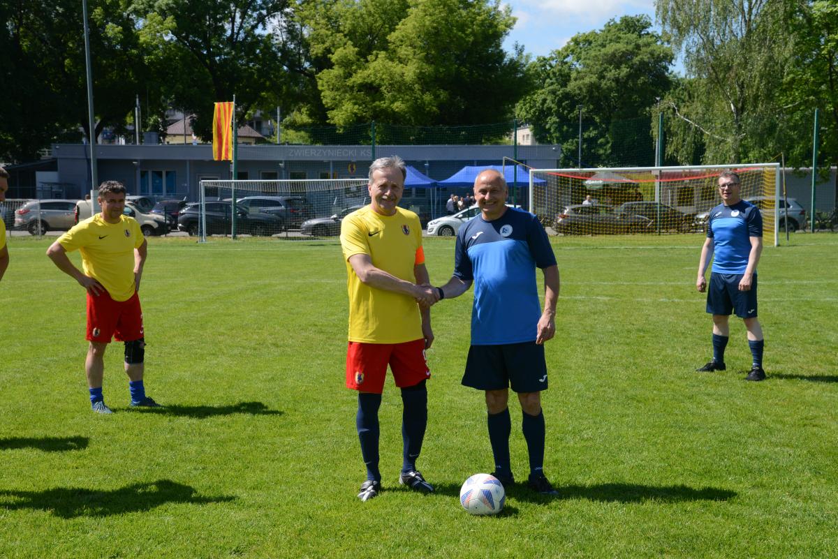 Zdjęcie nr: 9-DSC_3242 przedstawia czterech mężczyzn, dwóch ubranych w stój piłkarski koloru żółtoczerwonego a dwóch granatowoczarnego. Na pierwszym planie dwóch mężczyzn przeciwnych sobie drużyn podaje sobie ręce, pozostali dwaj się przyglądają. W tle znajdują się bramki piłkarskie.