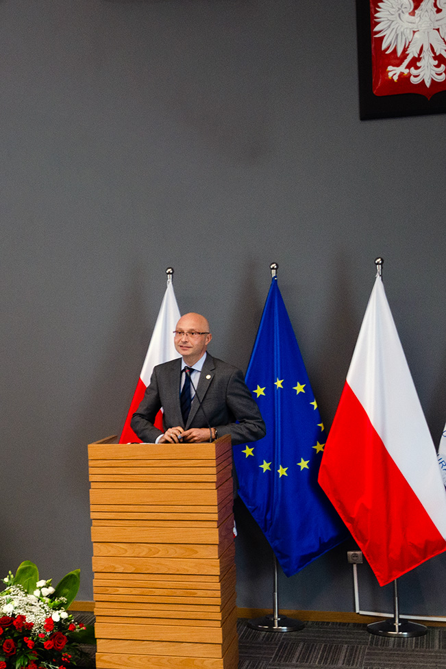 Przedstawia Dyrektora prof. dr. hab. Piotra Girdwoynia za mównicą. Za mężczyzną widać trzy flagi – dwie państwowe oraz unijną, umieszczone na stojakach flagowych. Na ścianie znajduje się godło państwowe. Przy mównicy, na podłodze leżą kwiaty w kolorze biało-czerwonym. Zdjęcie jest wykonane na tle szarej ściany. 