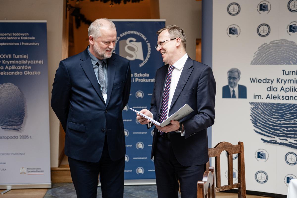 przedstawia wizerunki dwóch mężczyzn na tle rollup’ów z logo Instytutu Ekspertyz Sądowych im. dra Jana Sehna oraz Krajowej Szkoły Sądownictwa i Prokuratury oraz XXVII Turnieju Wiedzy Kryminalistycznej im. Aleksandra Głazka