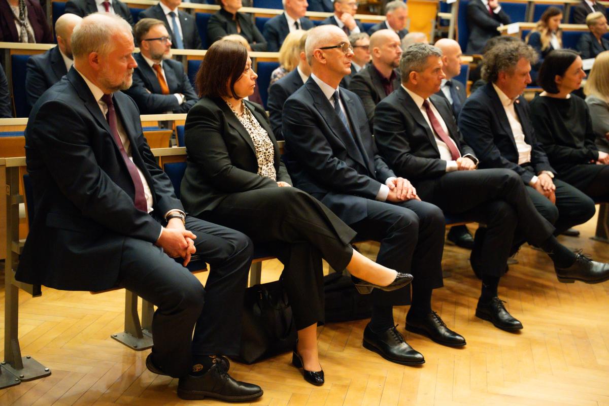 Widoczne osoby, kobiety i mężczyźni, w ciemnych garniturach, które siedzą w kilku rzędach na granatowych fotelach.