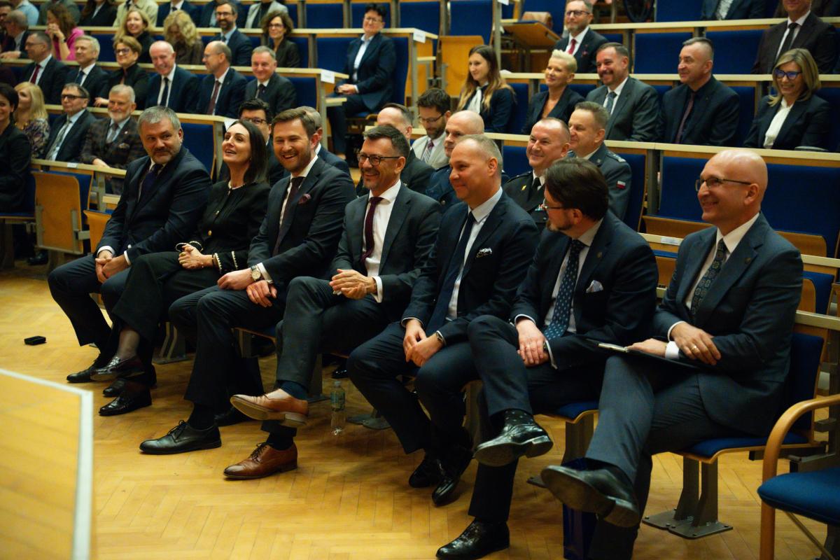 Wewnątrz auli widoczna grupa uśmiechniętych osób, kobiet i  mężczyzn, w ciemnych garniturach, którzy siedzą w kilku rzędach na granatowych fotelach.