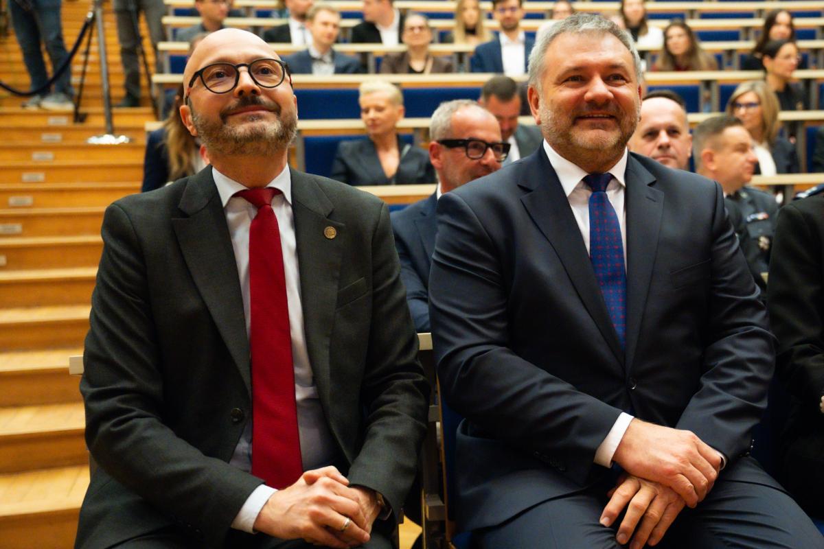 : Wewnątrz auli widocznych dwóch uśmiechniętych mężczyzn w ciemnych garniturach, białych koszulach i krawatach. W tle widoczne są osoby, kobiety i mężczyźni, które siedzą w kilku rzędach na granatowych fotelach.