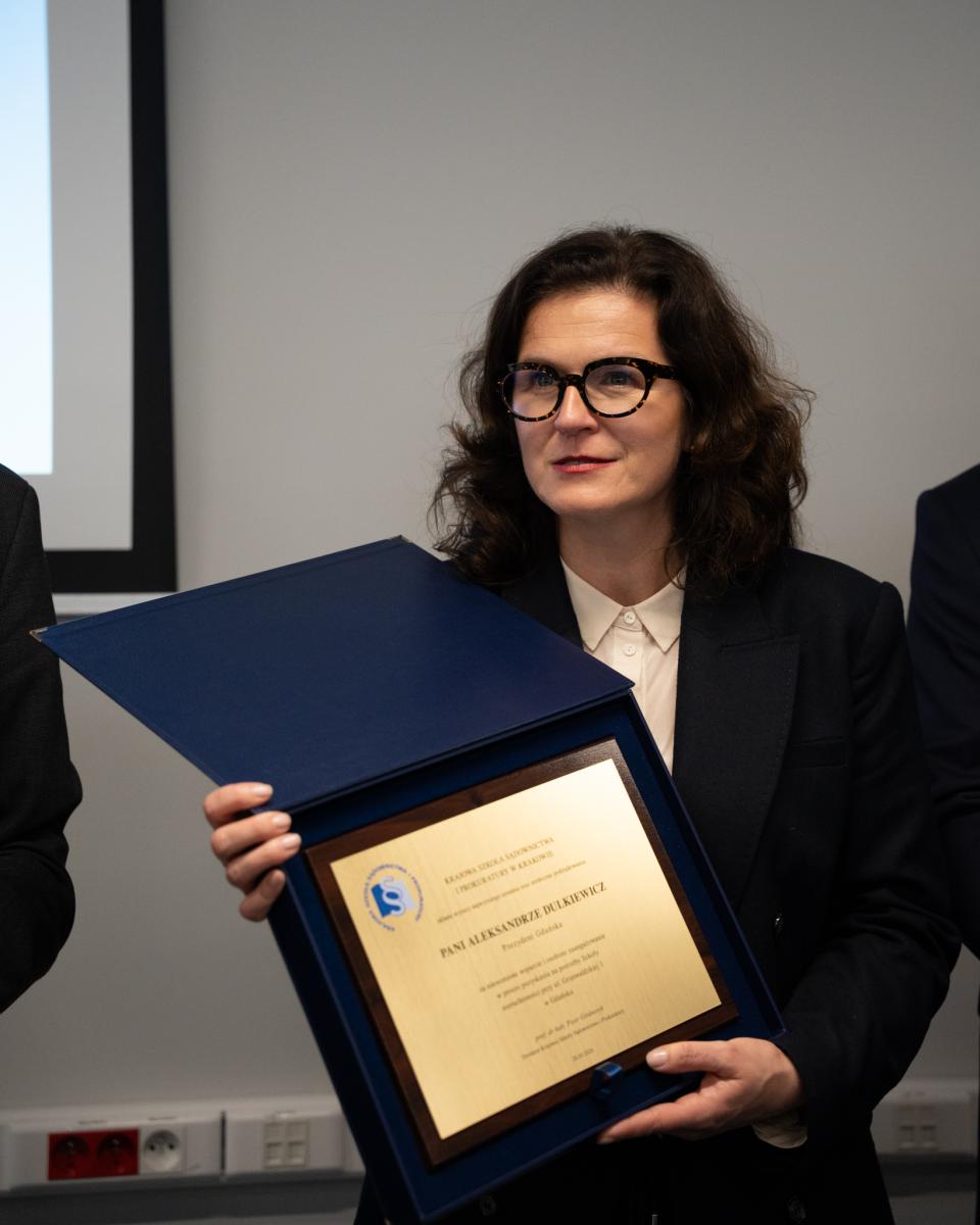 Widoczna postać kobiety w okularach, w ciemnej marynarce i białej bluzce na tle ściany. Kobieta trzyma w ręku obraz w brązowych ramkach w granatowej oprawie z tekstem oraz logo.  