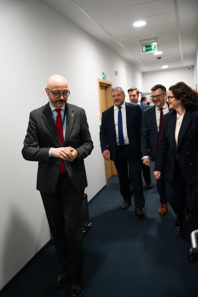 Widoczne postacie osób - czterech mężczyzn i jedna kobieta – którzy idą wąskim korytarzem. Osoby ubrane są w ciemne garnitury i białe koszule.
