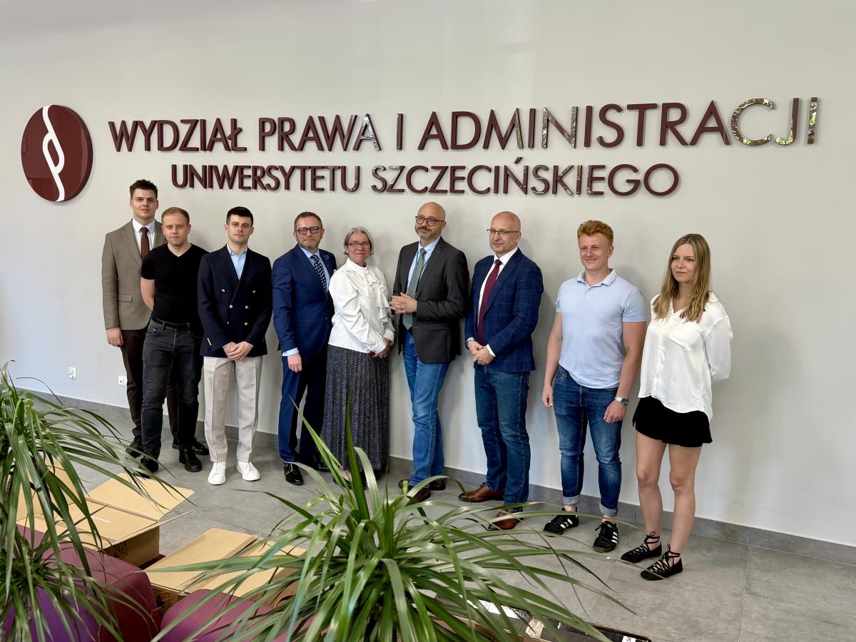 - na tle ściany z dużym napisem Wydział Prawa i Administracji Uniwersytetu Szczecińskiego stoją w rzędzie osoby pozujące do fotografii. Są to dwie kobiety ubrane w białe bluzki oraz siedmiu mężczyzn.