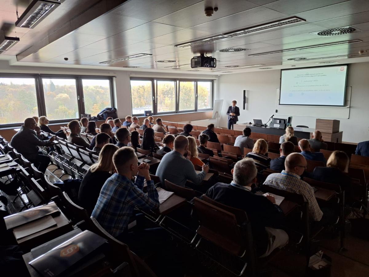 zdjęcie przedstawia  salę zajęciową  w trakcie zajęć , w tle sylwetka mężczyzny prowadzącego prelekcje , sylwetki uczestników zajęć widoczne od tyłu 