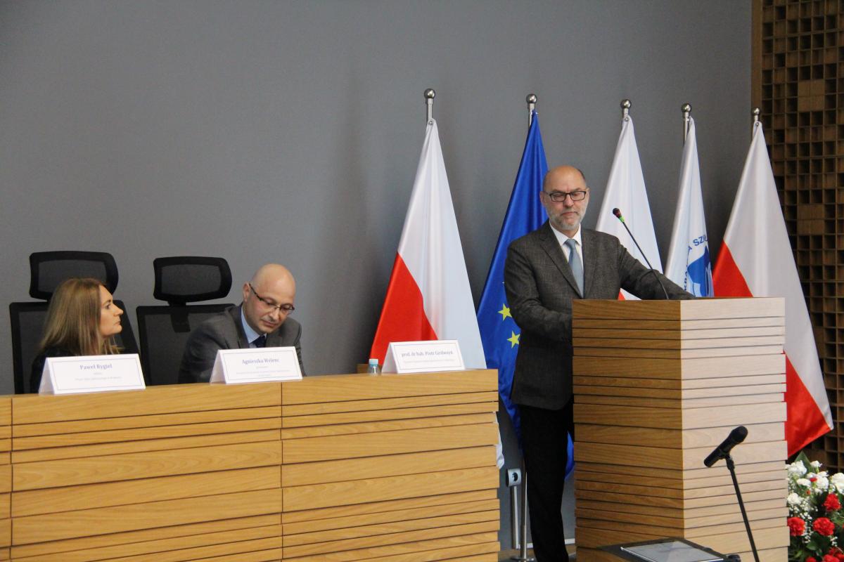 zdjęcie przedstawia przemawiającego mężczyznę w okularach i brodzie, na tle pięciu flag: białoczerwonej oraz flagi UE, obok za stołem z lewej strony siedzi kobieta i mężczyzna w okularach.