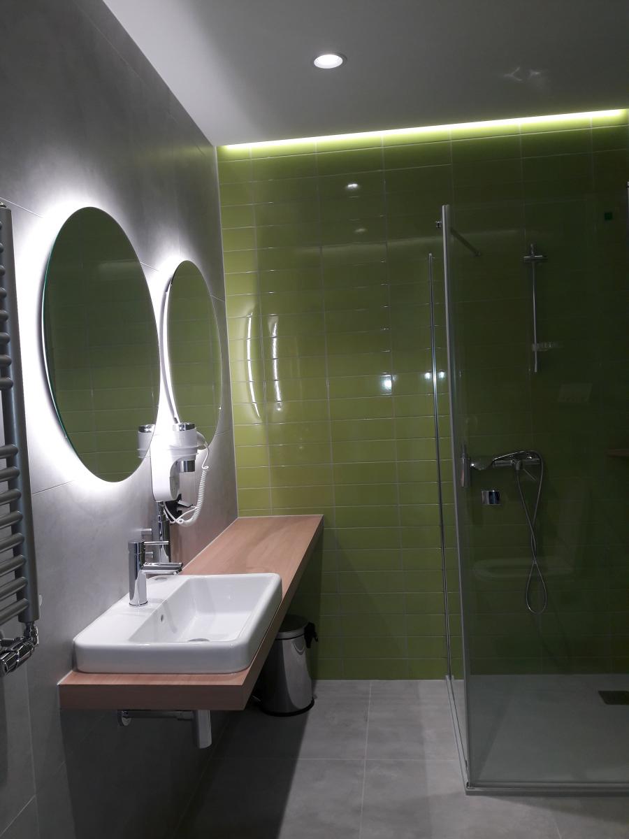 Załączone zdjęcie przedstawia łazienkę, znajdującą się w pokoju dwuosobowym z łóżkiem małżeńskim (typu DBL). Ściana po lewej stronie i podłoga pokryta jest płytkami koloru popielatego, natomiast ściana na wprost płytkami w kolorze zielonym. Po lewej stronie znajduje się długi blat łazienkowy wraz z wmontowaną umywalką i baterią łazienkową, pod blatem widoczny jest kosz na śmieci oraz syfon w kolorze srebrnym. Nad blatem do ściany przytwierdzony jest uchwyt na suszarkę do włosów w kolorze białym wraz z suszarką, a także dozownik do mydła. Nad blatem łazienkowym na ścianie wiszą dwa okrągłe, podświetlone lustra, które odbijają się w ścianie sąsiedniej. Obok lustra widoczny jest fragment grzejnika łazienkowego w kolorze popielatym. Po prawej stronie zdjęcia znajduje się kabina prysznicowa, przez szklane ściany kabiny można dostrzec srebrną baterię prysznicową wraz z drążkiem przymocowanym do ściany. Sufit podwieszany jest koloru białego, posiada półkę z oświetleniem LED, a na środku sufitu zamontowany jest także halogen.